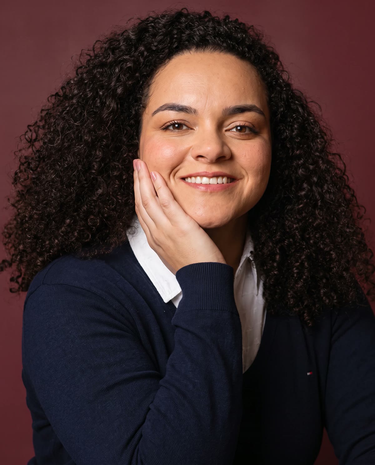 Tainá Motta com a mão no rosto, sorrindo, em fundo vinho